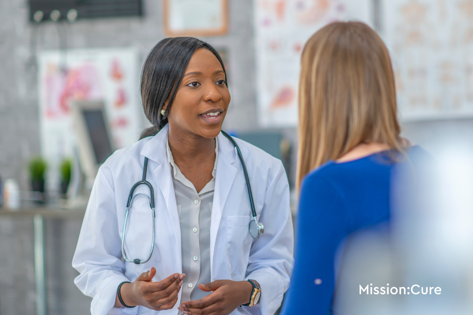 "Doctor in a white coat and stethoscope sitting and attentively speaking with a patient in a blue blouse during a medical consultation. The clinical setting includes anatomical charts and medical equipment in the blurred background. Mission logo is visible in the corner of the image.