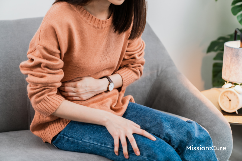 A person wearing a peach-colored sweater and jeans sits on a couch, clutching their abdomen in discomfort. The image conveys the experience of abdominal pain, which can be associated with pancreatitis or menstrual-related symptoms. The Mission: Cure logo is visible in the lower right corner of the image.